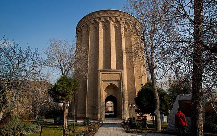A historic cylindrical tower made of brick, surrounded by trees, with a clear blue sky in the background.