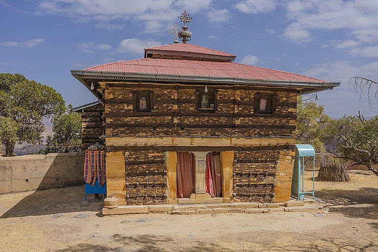 Exterior view of a traditional Ethiopian church built with wood and stone, featuring a cross on the roof and decorative elements.
