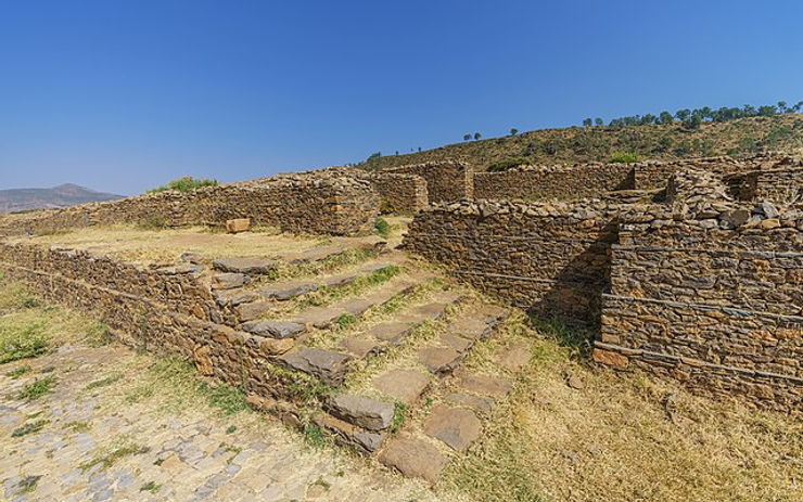 Ruins of ancient stone structures at a historical site, featuring steps and grassy areas under a clear blue sky.