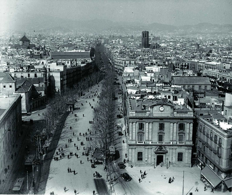 A historical black-and-white photograph of a bustling street in Barcelona, showing trees lining a wide avenue, people walking, and buildings in the background.