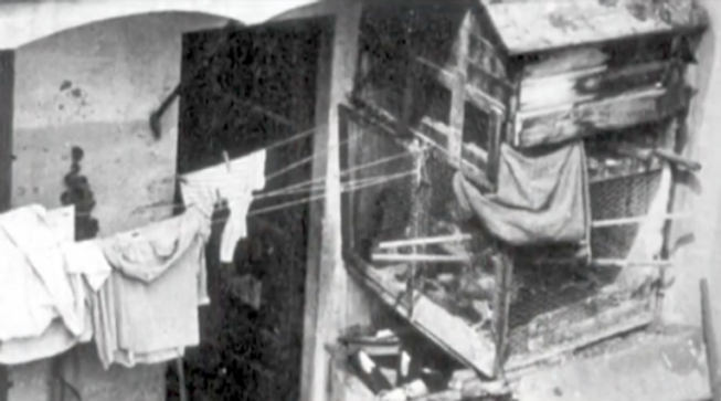 A black and white photograph depicting the exterior of a dilapidated building in El Raval, Barcelona, with laundry hanging outside and a makeshift structure attached to the wall.
