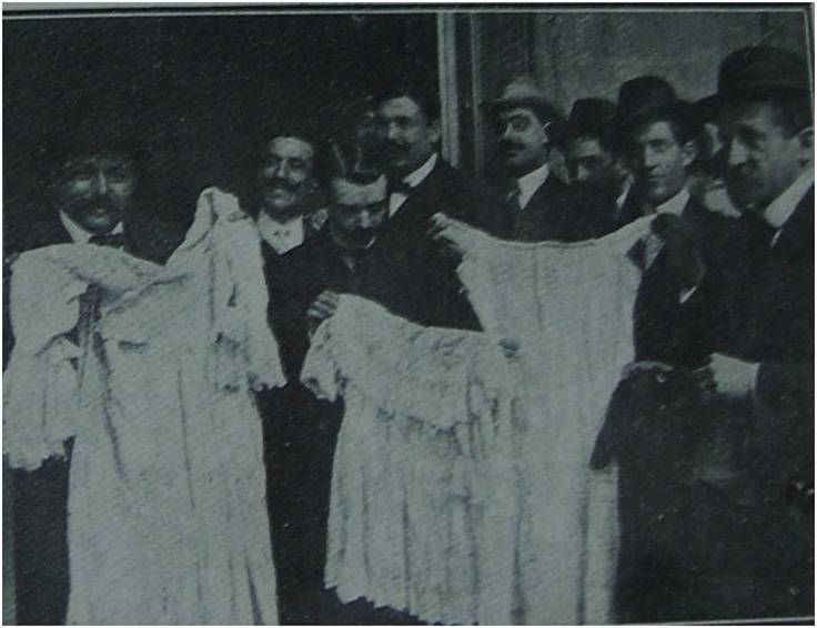 A historical black and white photograph of a group of men holding up two white garments, surrounded by others wearing hats and formal attire, possibly related to a criminal investigation in early 20th century Barcelona.