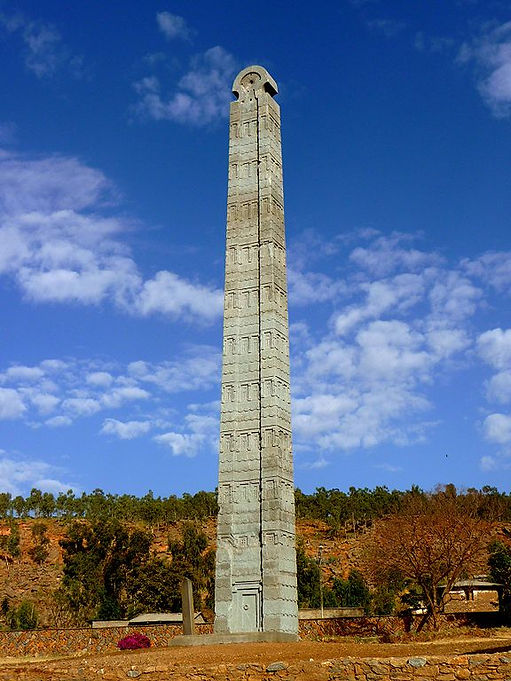 A large ancient obelisk from the Kingdom of Aksum, standing tall against a clear blue sky with scattered clouds, surrounded by a hillside with trees.
