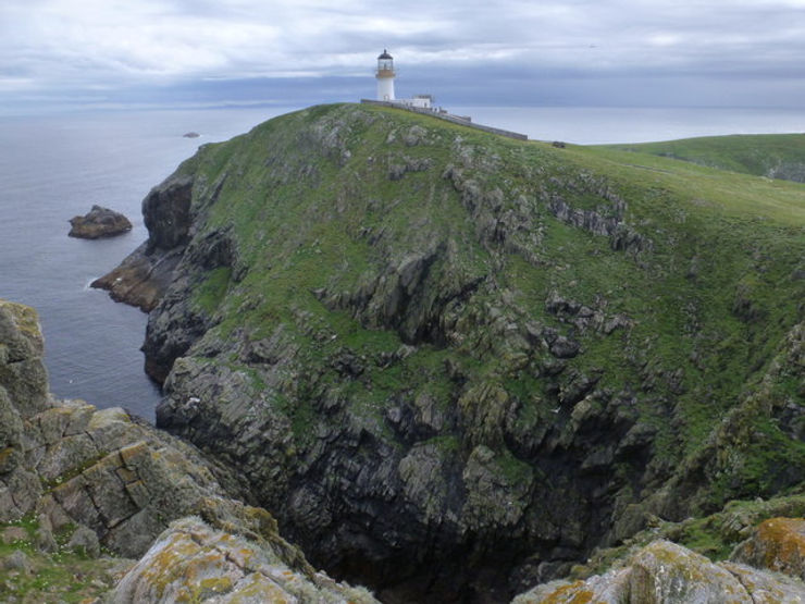 A panoramic view of Eilean Mòr, showing the Flannan Isles Lighthouse perched on a grassy cliff overlooking the ocean.