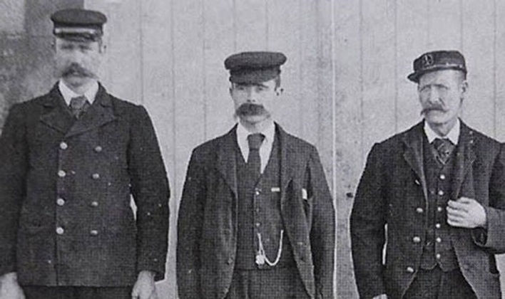 Three lighthouse keepers in historic uniforms standing in front of a wooden background.