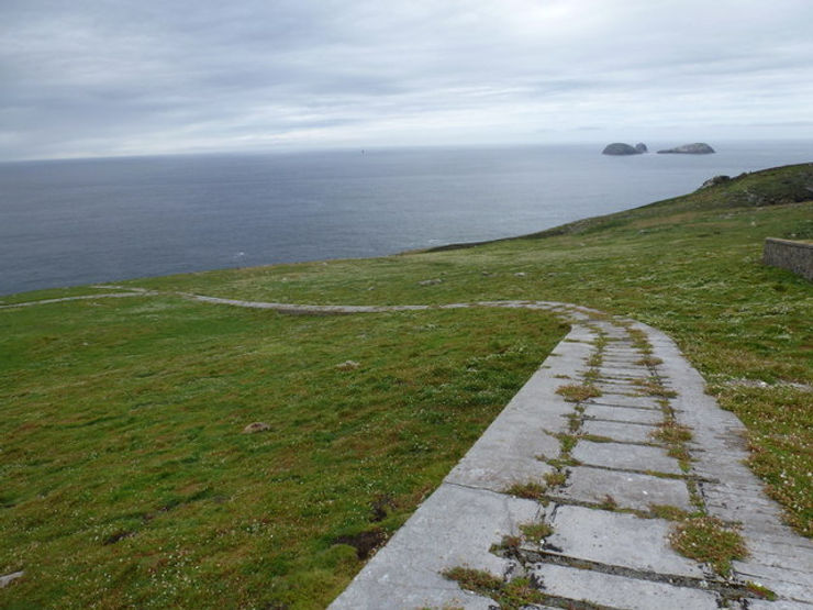 A grassy path leading towards the sea on Eilean Mòr, with distant islands visible in the background under a cloudy sky.