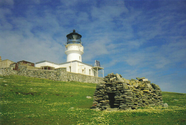A historic lighthouse on Eilean Mòr, surrounded by green grass and wildflowers, with an old stone chapel or structure in the foreground under a blue sky.