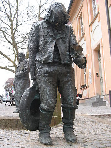 A bronze statue of Kaspar Hauser, depicting him in tattered clothing, holding a cup and a hat, with a downcast expression, situated in an outdoor setting.