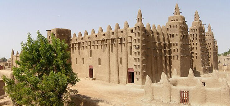 A view of the Great Mosque of Djenné, a historic adobe structure in Mali, showcasing its distinctive architectural features and surrounded by sandy terrain.
