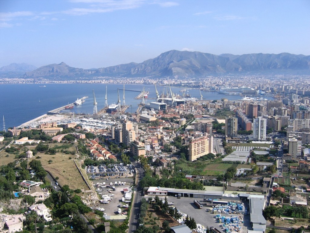 A panoramic view of Palermo, Italy, showcasing the port, cityscape, and surrounding mountains under a clear blue sky.